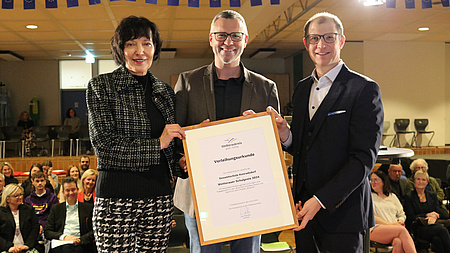 Schulleiterin Birgit Bingel, stellvertretender Schulleiter und Förderstufenleiter Christof Schranz und Landrat Jan Weckler stehen auf einer Bühne. Sie halten die Urkunde in die Kamera.