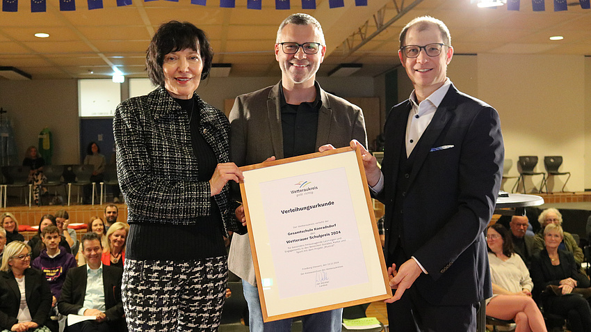 Schulleiterin Birgit Bingel, stellvertretender Schulleiter und Förderstufenleiter Christof Schranz und Landrat Jan Weckler stehen auf einer Bühne. Sie halten die Urkunde in die Kamera.