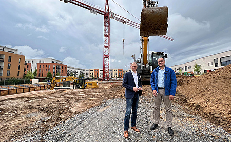 Zwei Männer auf einer Baustelle, rechts ein Sandhügel, im Hintergrund ein Kran.
