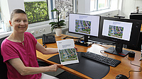 Eine Frau sitzt in einem Büro an einem Schreibtisch und hält ein Dokument in Händen. Die Grafik des Dokumententitels ist auch auf den Bildschirmen auf dem Schreibtisch zu sehen.