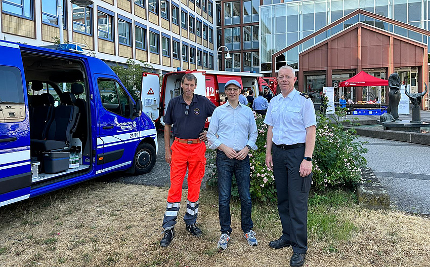 Drei Männer stehen auf dem Platz vor einem Gebäude. Daneben und im Hintergrund Fahrzeuge des Rettungsdienstes und der Feuerwehr.