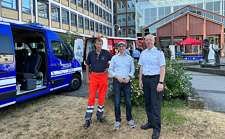 Drei Männer stehen auf dem Platz vor einem Gebäude. Daneben und im Hintergrund Fahrzeuge des Rettungsdienstes und der Feuerwehr.