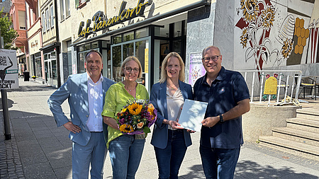 Vier Personen stehen in einer Fußgängerzone zusammen. Zwei Frauen und zwei Männer. Die eine Frau übergibt einen Förderbescheid an einen der Männer, die andere Frau hält einen Blumenstrauß in den Händen