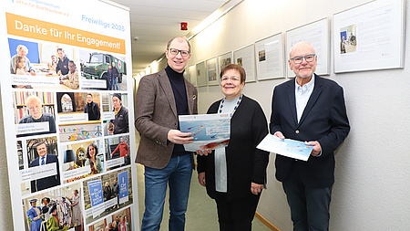 Ein Frau und zwei Männer stehen gemeinsam in einem Flur. Die Männer halten jeweils Wandkalender in Händen. Im Hintergrund hängen mehrere Bilderrahmen an der Wand. Links von der Gruppe steht ein Plakat.