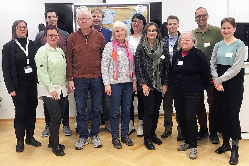 Eine kleine Personengruppe von Männern und Frauen. Die Personen stehen neben- und hintereinander. Im Hintergrund Pinwände.