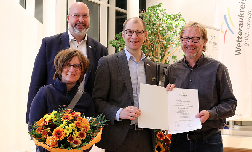 Drei Männer und eine Frau stehen beieinander. Zwei der Männer halten gemeinsam eine Urkunde, die Frau hält einen Blumenstrauß in der Hand