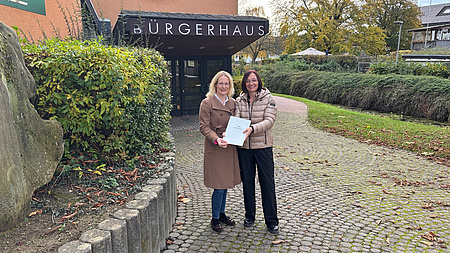 Zwei Frauen stehen vor einem Bürgerhaus und übergeben eine Mappe mit einem Zuwendungsbescheid.