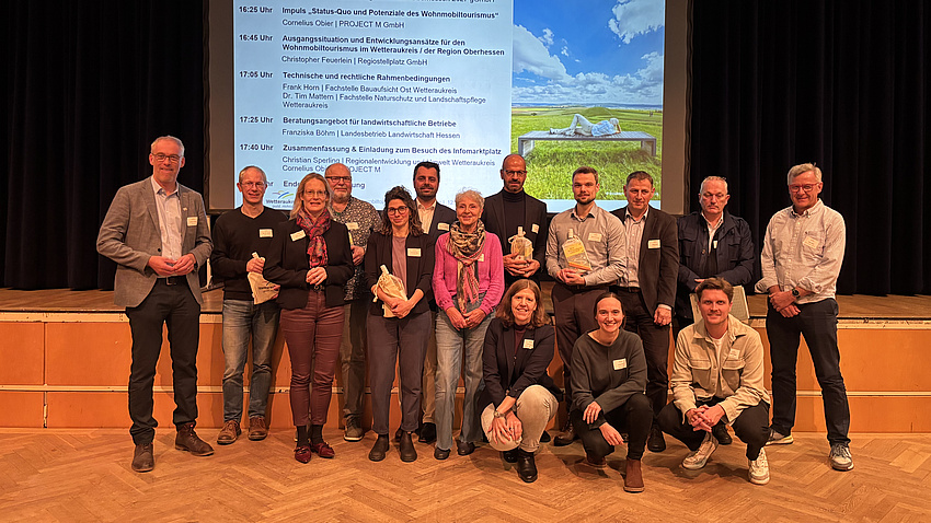 Eine größere Gruppe von Menschen posiert für das Foto vor einer Tribüne anlässlich der Veranstaltung zum Wohnmobiltourismus im Wetteraukreis. die meisten Personen stehen, einige knien davor.