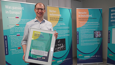 Ein Mann steht vor großen Plakaten und hält einen Bilderrahmen in den Händen.