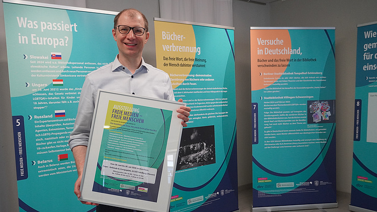 Ein Mann steht vor großen Plakaten und hält einen Bilderrahmen in den Händen.