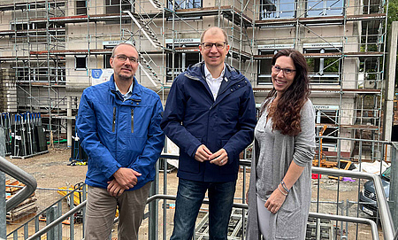 Zwei Männer und eine Frau stehen auf einem Balkon. Im Hintergrund ein eingerüstetes Gebäude, das sich noch im Rohbau befindet.