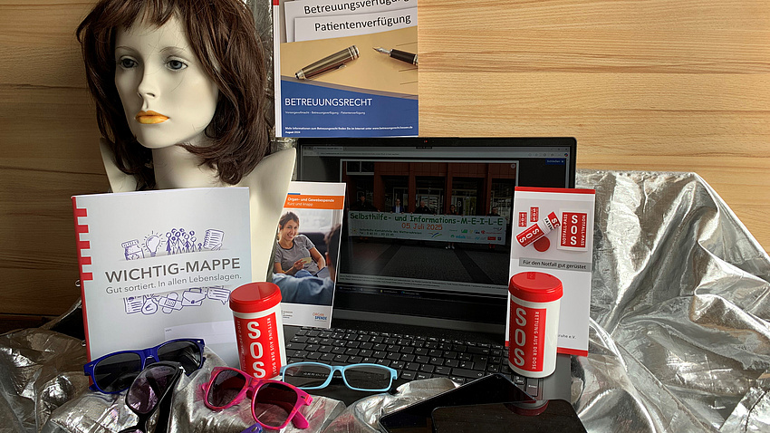 Auf dem Bild sieht man alles, was an der Selbsthilfe-MEiLE verteilt bzw. gesammelt wird: Perücken, SOS,Dose, Brillen, Handys und Tablets, Wichtigmappen und Betreuungs- sowie Vorsorgebroschüren.