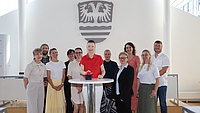 Mehrere Frauen und Männer stehen in einem Saal um einen Tisch. Darauf befindet sich ein großer, roter Knopf, den eine der Frauen drückt.