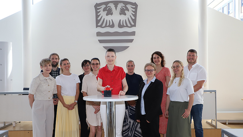 Mehrere Frauen und Männer stehen in einem Saal um einen Tisch. Darauf befindet sich ein großer, roter Knopf, den eine der Frauen drückt.