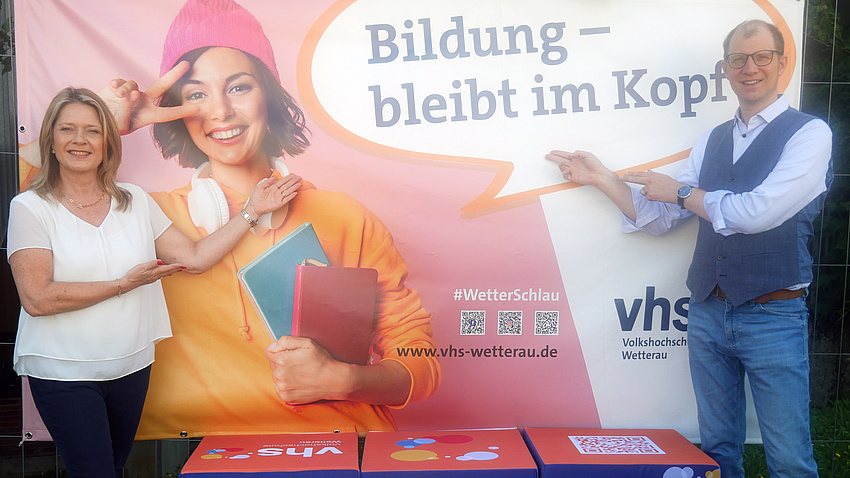 Eine Frau und ein Mann stehen vor einer Plakatwand und zeigen auf diese.