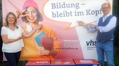 Eine Frau und ein Mann stehen vor einer Plakatwand und zeigen auf diese.