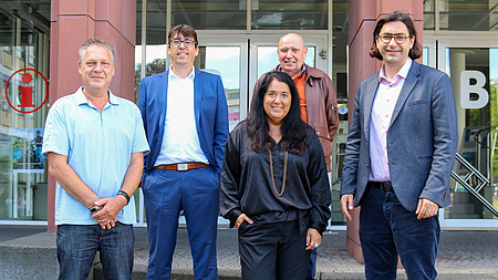 Vier Männer und eine Frau stehen vor einem Gebäude. Sie lächeln in die Kamera.