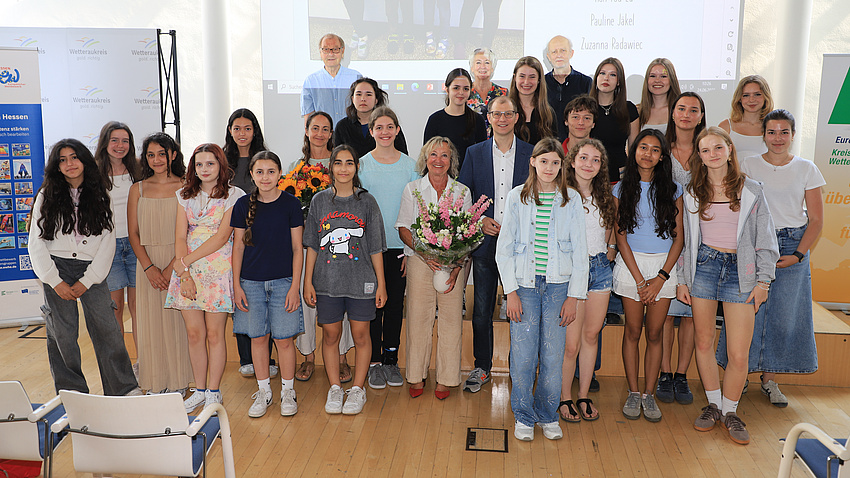 Zahlreiche Jugendliche und fünf Erwachsene stehen in einem Saal vor einer Leinwand.