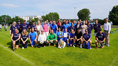 Zahlreiche Frauen und Männer, ein großer Teil von ihnen im Fußball-Trikots, stehen oder knien auf dem Rasen eines Sportfelds. In ihrem Mittelpunkt befindet sich ein Pokal.