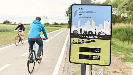 Zwei Radfahrer sind auf einem Radweg unterwegs. Neben dem Radweg steht ein Display, auf dem "Fahrradzählstation" und Zahlen zu lesen sind.