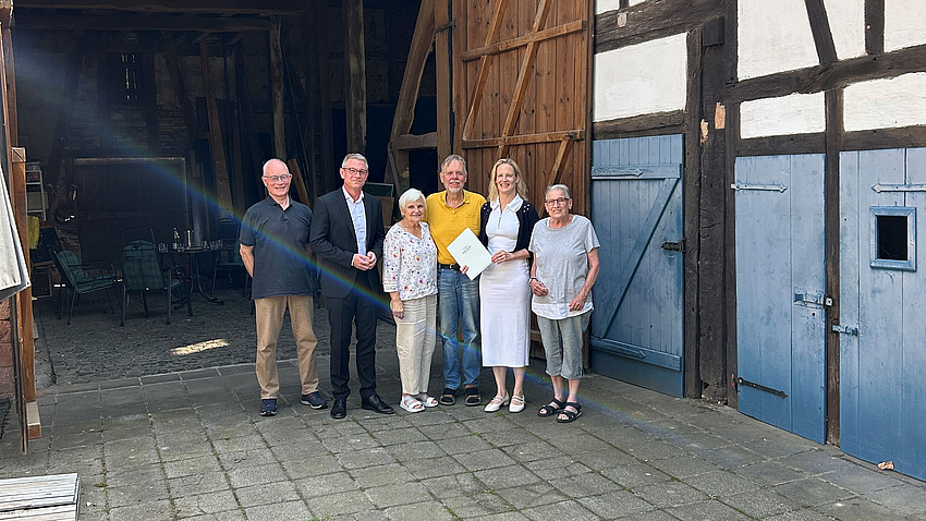 Drei Frauen und drei Männer stehen in einer Hofeinfahrt vor einem geöffneten Scheunentor. Angrenzend ist eine Fachwerkwand zu sehen. Eine der Frauen hält eine Urkunde in Händen.