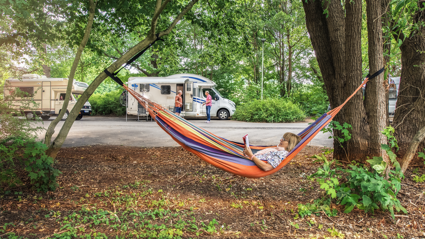 Eine Person liegt auf einer Hängematte und liest ein Buch, im Hintergrund sind mehrere Wohnmobile zu sehen.