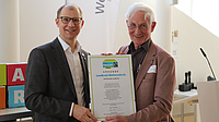 Zwei Männer halten in einem Saal gemeinsam eine Urkunde in der Hand.