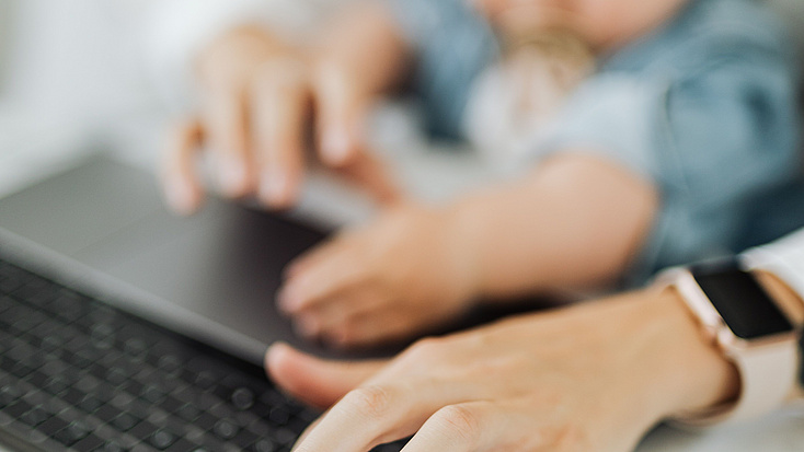 Das Foto zeigt verschwommen eine Kleinkind auf dem Schoß eines Elternteils vor eine Laptop. Es ist ein Symbolbild für die Vereinbarkeit von Familie und Beruf.