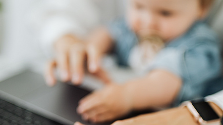Das Foto zeigt verschwommen eine Kleinkind auf dem Schoß eines Elternteils vor eine Laptop. Es ist ein Symbolbild für die Vereinbarkeit von Familie und Beruf.