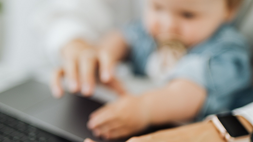 Das Foto zeigt verschwommen eine Kleinkind auf dem Schoß eines Elternteils vor eine Laptop. Es ist ein Symbolbild für die Vereinbarkeit von Familie und Beruf.
