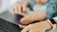 Das Foto zeigt verschwommen eine Kleinkind auf dem Schoß eines Elternteils vor eine Laptop. Es ist ein Symbolbild für die Vereinbarkeit von Familie und Beruf.