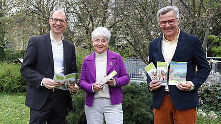 Zwei Männer und eine Frau stehen vor einem blühenden Baum. Sie halten eine Broschüre in die Kamera.