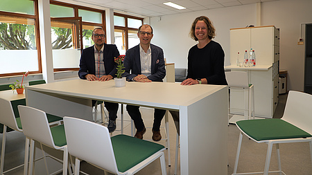 Zwei Männer und eine Frau sitzen auf modernen Möbeln und lächeln in die Kamera.