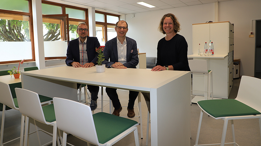 Zwei Männer und eine Frau sitzen auf modernen Möbeln und lächeln in die Kamera.