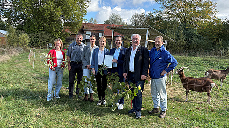 Drei Frauen und vier Männer stehen gemeinsam auf einer Wiese. Im Hintergrund sind auf einer abgezäunten Weidefläche Ziegen zu sehen und dahinter das Dach eines Gebäudes. Eine der Frauen und einer der Männer halten gemeinsam ein Dokument in Händen.