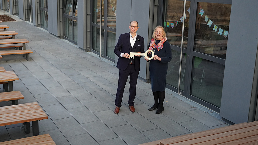 Landrat Jan Weckler und Schulleiterin Cornelia Jüttner-Tunkowski stehen vor einem Gebäude. Sie halten einen großen goldenen Schlüssel in Händen.
