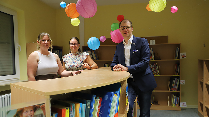 Zwei Frauen und ein Mann stehen in einem Raum an einem Buchregal. Im Hintergrund sind weitere Regale zu erkennen und von der Decke hängen mehrere bunte Papier-Lampions.