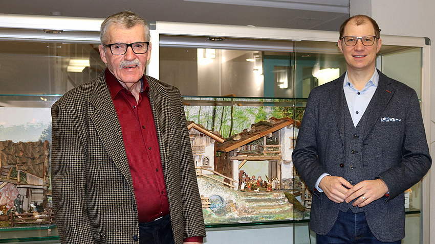 Zwei Männer stehen vor einer Glasvitrine, in der eine Weihnachtskrippe ausgestellt ist.