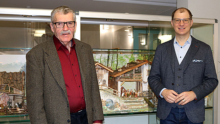 Zwei Männer stehen vor einer Glasvitrine, in der eine Weihnachtskrippe ausgestellt ist.