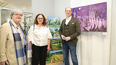 Zwei Frauen und ein Mann stehen in einem größeren Raum vor mehreren Landschaftsgemälden.