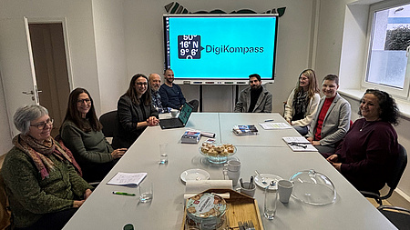 Sechs Frauen und drei Männer sitzen an einem Tisch in einem größeren Raum. Im Hintergrund befindet sich ein Bildschirm an der Wand.