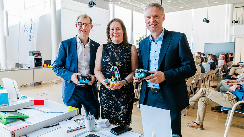 Landrat Jan Weckler, Fachdienstleiterin Katja Müller-Lind und Staatssekretär Dr. Manuel Lösel stehen im Plenarsaal. Sie halten kleine Roboter aus in der Hand.