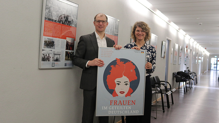 Der Landrat und die Frauenbeauftragte stehen mit einem Plakat in der Hand vor der Plakatausstellung und schauen in die Kamera.