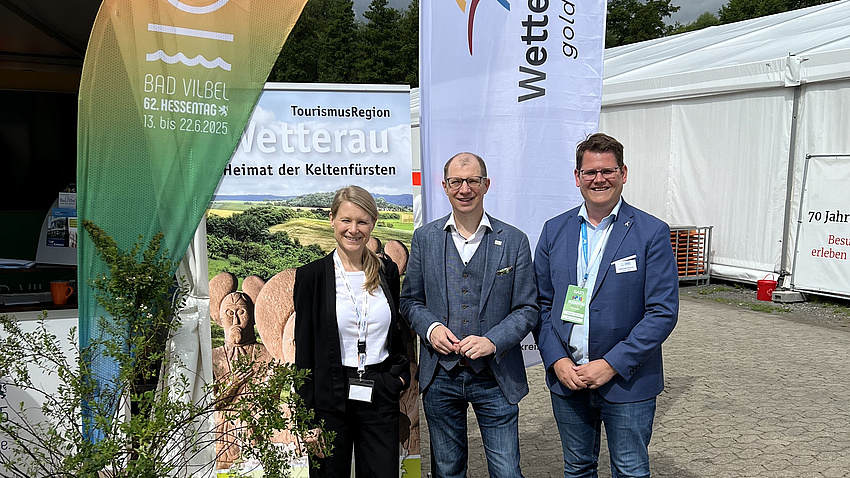 Landrat Jan Weckler (Mitte), Bürgermeister Sebastian Wysocki und Hessentagskoordinatorin Anne Kemink. Sie lächeln in die Kamera. Im Hintergrund Fahnen der Stadt Bad Vilbel und des Wetteraukreises.