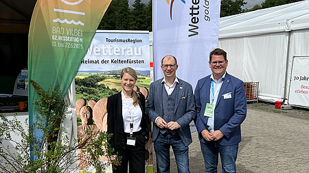 Landrat Jan Weckler (Mitte), Bürgermeister Sebastian Wysocki und Hessentagskoordinatorin Anne Kemink. Sie lächeln in die Kamera. Im Hintergrund Fahnen der Stadt Bad Vilbel und des Wetteraukreises.