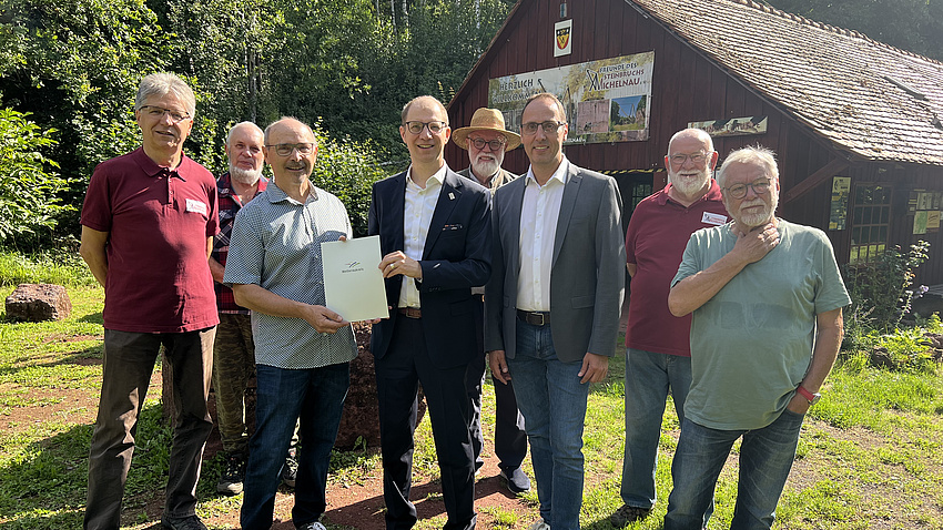 Acht Männer stehen auf einem Platz vor einem Gebäude, im Hintergrund sind Pflanzen zu sehen. Zwei der Männer halten zusammen eine Urkunde in Händen.