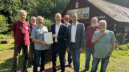 Acht Männer stehen auf einem Platz vor einem Gebäude, im Hintergrund sind Pflanzen zu sehen. Zwei der Männer halten zusammen eine Urkunde in Händen.