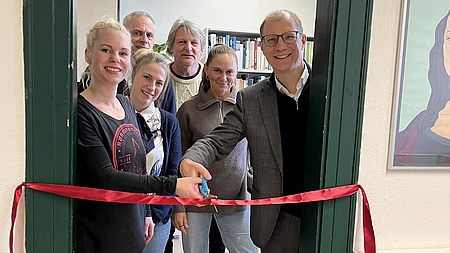 Mehrere Personen stehen in einem Türrahmen, der Landrat und eine Frau durchschneiden ein Band.