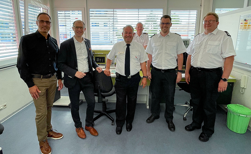 Fünf Männer stehen vor einem Arbeitstisch mit technischen Geräten. Im Hintergrund ein weiterer Mann.