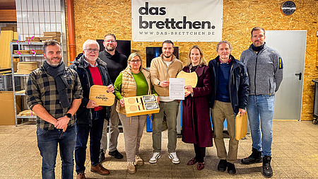 Zwei Frauen und sechs Männer stehen gemeinsam in einer größeren Betriebshalle. Eine der Frauen und einer der Männer halten gemeinsam ein Dokument in Händen.